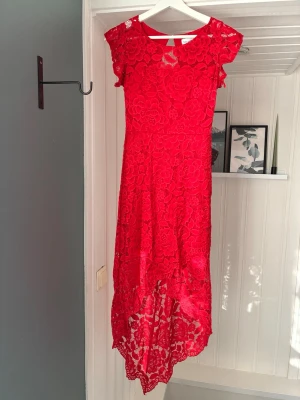 Röd spetsklänning från Mix by Heart - Säljer en röd klänning från Mix by Heart med vacker spets och blommigt mönster. Klänningen har korta ärmar, rund halsringning och asymmetrisk nederkant som är längre bak. Perfekt för dig som vill sticka ut med en färgstark look.