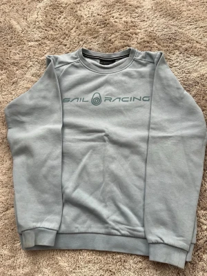Ljusgrå Sail Racing sweatshirt - Ljusgrå sweatshirt från Sail Racing med tryckt logga på bröstet och liten logga bak. Tröjan har rund hals, ribbade muddar och är tillverkad i mjukt bomullsmaterial. Perfekt för en avslappnad och sportig stil. OBS! Barnstorlek 150