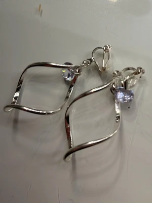 Silvriga örhängen med lila kristall - Unika örhängen i silverfärgad metall med böljande, twistad form och en hängande lila kristall. Clip-on fäste gör dem enkla att använda även utan hål i öronen. Perfekt för dig som gillar stilrena och moderna accessoarer med en touch av färg.