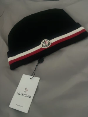 Svart Moncler mössa med röd-vit rand - Snygg svart mössa från Moncler med klassisk röd och vit rand runt om samt Moncler-logga framtill. Mössan har en enkel, stilren design och är tillverkad i mjukt material som sitter skönt på huvudet. Perfekt accessoar för dig som gillar streetwear och exklusiva märken.