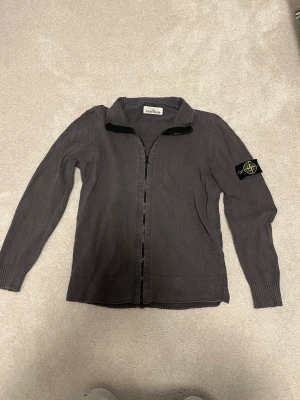 Grå zip-kofta från Stone Island - Säljer en grå stickad kofta från Stone Island med hel dragkedja och klassisk logga på ärmen. Tröjan har lång ärm, ribbade muddar och en enkel krage. Perfekt för lager-på-lager och snygg till jeans eller cargobyxor. Pris kan diskuteras vid snabbaffär!