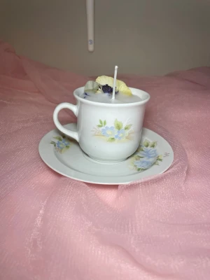 Doftljus i vaniljdoft i en tekopp med fat i blommig design  - Det här ljuset är gjort av återanvändbara material. Tekoppen är köpt secondhand och stearinet är från kyrkor som tänkte slänga det. Det här ljuset har en svag doft utav vanilj, så känsliga även kan använda det. Det här ljuset har även torkade blommor på som dekoration. 