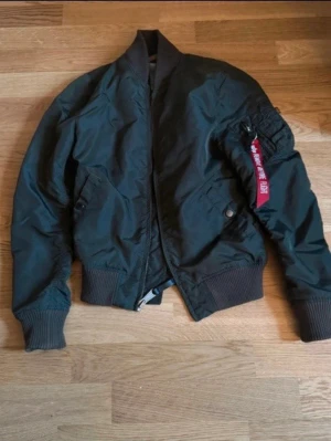 Mörkgrön bomberjacka Alpha Industries - Säljer en klassisk mörkgrön bomberjacka från Alpha Industries med ikonisk röd 'Remove Before Flight'-tag på ärmen. Den är i väldigt bra skickar bara använts några gånger