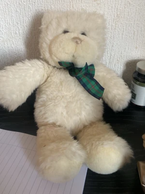 Vit mjuk nallebjörn med rosett - Söt vit nallebjörn med fluffig päls och en grönblå-rutig rosett runt halsen. Nallen har rund nos, små svarta ögon och mjuka former. Perfekt som dekoration eller present till någon speciell.