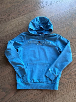 Blå hoodie från Tommy Hilfiger - Snygg blå hoodie från Tommy Hilfiger med broderad logga på bröstet Klassisk känguruficka framtill och ribbade muddar vid ärmslut och nederkant. Tillverkad i mjukt bomullsmaterial och har en bekväm huva.