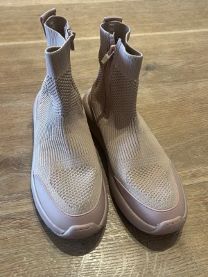 Beige stickade sneakers från Zara - Säljer ett par beige stickade sneakers från Zara med högt skaft och dragkedja på sidan. Skorna har en chunky sula i gummi och ovandel i stretchigt stickat material som ger en modern och sportig look.