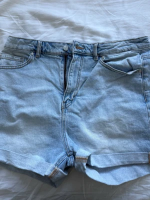 Ljusblå denimshorts med hög midja - Säljer ett par ljusblå loose fit denimshorts med hög midja och uppvikta benslut. Klassisk femficksmodell med dragkedja och knapp framtill. Perfekta till sommaren och lätta att matcha med olika stilar.