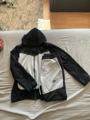 Nike svart och grått loungewear-set - Nike loungewear-set med zip-hoodie och matchande byxor i svart och ljusgrått. Hoodien har dragkedja, huva och en unik detaljficka på ärmen. Byxorna har resår i midjan och sidofickor. Perfekt för chill dagar hemma eller när du vill vara bekväm.