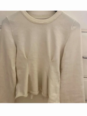 Beige långärmad tröja med volang - Säljer en beige långärmad tröja med rund hals och volangdetalj nedtill. Tröjan har en enkel och stilren design, perfekt för dig som gillar en clean look. Materialet känns mjukt och bekvämt mot huden.