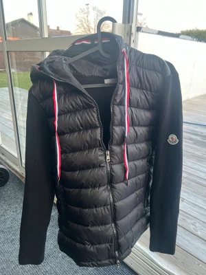 Svart Moncler cardiganjacka med huva - Snygg svart cardiganjacka från Moncler med vadderad front och stickade ärmar. Jackan har huva med röda och vita dragsnören samt Moncler-logga på ärmen. Dragkedja framtill och två sidofickor. Perfekt för lager-på-lager och streetstyle.