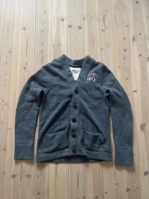 Grå kofta från Hollister - Mysig grå kofta från Hollister med knappar framtill och två fickor. Broderad logga och text '1922' på bröstet. Koftan har ribbade muddar vid ärmslut och en klassisk sjalkrage. Perfekt för lager på lager och chill stil.