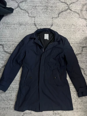 Mörkblå rock från J.Lindeberg - Stilren mörkblå rock från J.Lindeberg med klassisk krage och dold dragkedja. Rocken har två fickor med knapp framtill och är helfodrad. Perfekt för dig som vill ha en enkel och snygg look till hösten eller vintern.