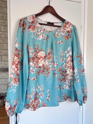 Blommig turkos blus, storlek L - Turkos blus med blommigt mönster i rostrött, vitt och brunt. Långa ärmar med resår vid ärmslut. Luftig och skön, använd 1 gång.