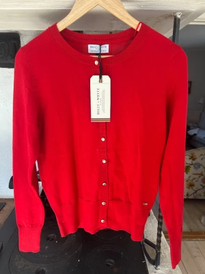 Röd kofta från Holly & Whyte - Snygg röd kofta från Holly & Whyte med rund halsringning och knappar framtill. Klassisk och enkel design med ribbade muddar vid ärmslut och nederkant. Perfekt att slänga över en t-shirt eller blus för en stilren look.