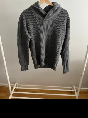 Grå stickad hoodie med krage - Säljer en grå stickad hoodie med bred ribbstickning och unik krage med knappdetalj. Tröjan har långa ärmar och en avslappnad passform, perfekt för kalla dagar. Materialet känns mjukt och värmande.