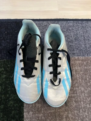 Adidas Messi fotbollsskor blå/vit - Snygga Adidas Messi fotbollsskor i vitt med ljusblå detaljer och svarta skosnören. Skorna har en låg profil och är tillverkade i syntetmaterial för bra bollkontroll. Perfekta för dig som vill sticka ut på planen med stilrena och sportiga skor.