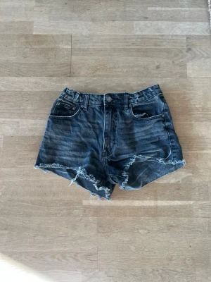 Svarta jeansshorts med fransig kant - Säljer ett par svarta jeansshorts med rå, fransig kant och klassisk femficksmodell. Shortsen har hög midja och slitna detaljer för en avslappnad look. Perfekta till sommaren och lätta att matcha med olika stilar.