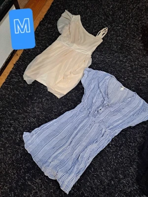 Två korta sommarklänningar - Säljer två korta klänningar, en i ljusbeige med oneshoulder och smala axelband, samt en blåvit randig med v-ringning och knappar framtill. Båda har en luftig passform och är perfekta för varma dagar.