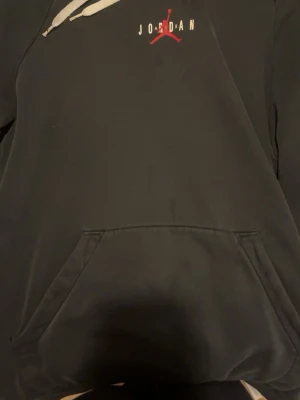 Svart Jordan hoodie med röd logga - Snygg svart hoodie från Jordan med den klassiska röda Jumpman-loggan och texten 'JORDAN' på bröstet. Hoodien har vita dragsnören, känguruficka framtill och en röd detalj på ärmen. Perfekt för en sportig och avslappnad stil.