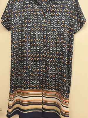 Mönstrad blus från Mango - Snygg blus från Mango med korta ärmar och grafiskt mönster i marinblått, vitt och orange. Blusen har en rak passform och dekorativa ränder i beige, brun och vit längst ner. Tillverkad i ett mjukt, lätt material som känns skönt mot huden.