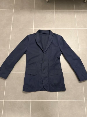 Mörkblå Kavaj från Raplh Lauren, stl M - Stilren mörkblå kavaj i bomull med tre knappar framtill, klassisk krage och tre utanpåliggande fickor. Perfekt för en avslappnad men ändå uppklädd look. Passar till både jeans och chinos.