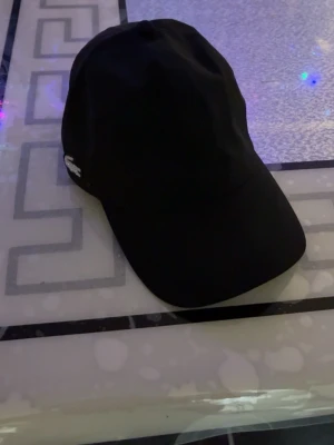 Svart keps från Lacoste - Snygg svart keps från Lacoste med den klassiska krokodilloggan broderad i vitt på sidan. Kepsen har böjd skärm och justerbar rem bak. Tillverkad i ett mjukt och slitstarkt material som passar perfekt till streetwear och casual outfits. Köpt för 1000kr 