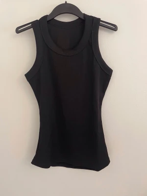 Svart ribbad linnetopp - Säljer en svart ribbad linnetopp med breda axelband och rund halsringning. Toppen är tight och ärmlös, perfekt att matcha med jeans eller kjol. Enkel och stilren design som passar till många olika outfits.
