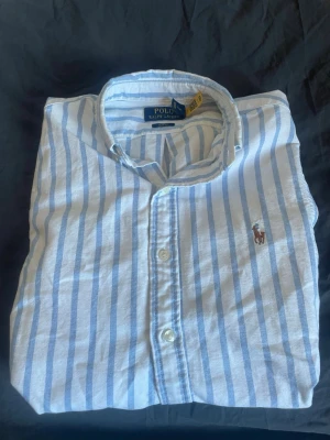Randig skjorta från Polo Ralph Lauren - Klassisk vit skjorta med ljusblå ränder från Polo Ralph Lauren. Skjortan har lång ärm, knappar framtill och den ikoniska broderade loggan på bröstet. Tillverkad i mjuk bomull och perfekt för en clean och stilren look. Köptes förra året för 2700 kr. Använd ca 3 ggr så i mycket fint skick.  Först till kvarn (för billigt)