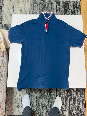 Blå kortärmad skjorta med krage - Snygg blå skjorta med korta ärmar och klassisk krage med röd och vit kant. Skjortan har knappar framtill och en bröstficka. Perfekt för dig som gillar. en sportig och stilren look.