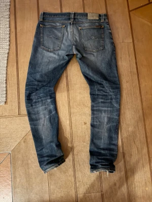 Nudie jeans - Säljer ett par blå skinny jeans med slitningar och hål på vänster knä. Jeansen har klassisk femficksdesign, låg midja och dragkedja. Materialet är denim i bomull med en tvättad look och smal passform. De har slitnings över hela och ett hål samt lagning på höger knä för en bättre look. W31 L32
