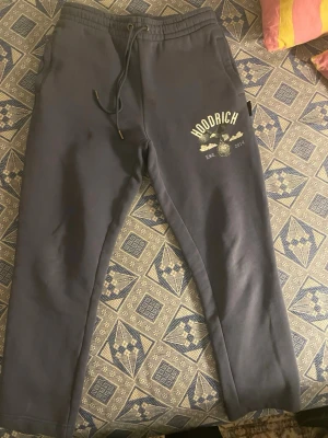 Mörkblå mjukisbyxor från Hoodrich - Mörkblå mjukisbyxor från Hoodrich med tryck på ena benet som visar en örn, texten 'HOODRICH' och 'EST. 2014'. Byxorna har dragsko i midjan och raka ben. Perfekta för en avslappnad och street-inspirerad stil.