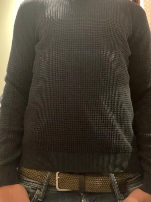 Mörkblå stickad tröja - En enkel och stilren mörkblå stickad tröja med diskret rutmönster. Tröjan har rund halsringning, långa ärmar och ribbade muddar vid ärmslut och nederkant. Perfekt att matcha med jeans för en avslappnad look.