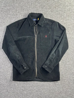 Polo Ralph Lauren Corduroy Overshirt – Size S - Corduroy overshirt från Polo Ralph Lauren i mörk färg med klassisk broderad pony-logga på bröstet. Tidlös modell som funkar perfekt som både skjorta eller lätt jacka.  📏 Storlek: S 📐 Mått visas i bilderna ✨ Skick: Bra skick – se bilder för detaljer  Perfekt för preppy, casual eller vintage outfits.  💸 Följ mig så får du 10% rabatt på köpet!