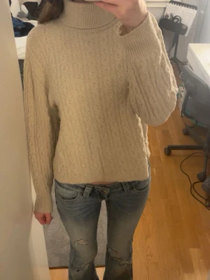 Beige polotröja med kabelstickning - Mysig beige stickad polotröja med klassiskt kabelstickat mönster. Tröjan har hög polokrage, långa ärmar och ribbade muddar vid ärmslut och nederkant. Perfekt för kyliga dagar och enkel att matcha med jeans eller kjol.