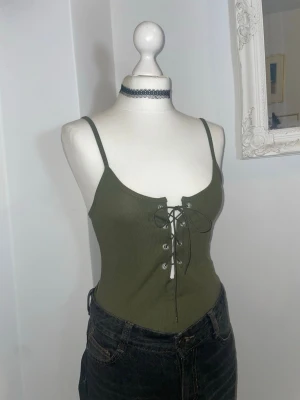 Olivgrön ribbad body med snörning - Olivgrön ribbad body med smala axelband och snygg snörning framtill. Toppen har en tight passform. Köpt i London.
