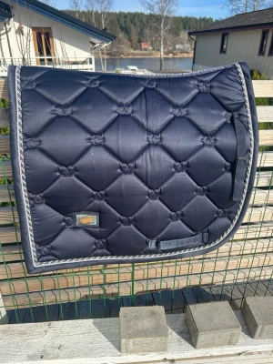 Mörkblått schabrak från equestrian Stockholm - Snyggt mörkblått dressyr schabrak med quiltat mönster och dekorativ silverfärgad kant. Passar perfekt till ridning och ger en stilren look till din hästutrustning. Fästen för sadel och gjord finns.