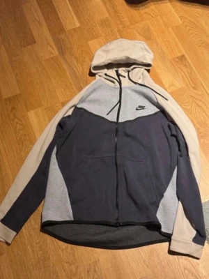 Nike hoodie i grå och beige färgblock - Nike hoodie med dragkedja i färgblockad design med mörkgrå, ljusgrå och beige. Klassisk huva med dragsko och två fickor framtill. Tillverkad i mjukt bomullsmaterial, perfekt för chill dagar. Nike-logga på bröstet för en sportig vibe.