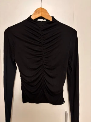 Svart långärmad topp från Denim Tom Tailor - Säljer en svart långärmad topp från Denim Tom Tailor. Toppen har en hög hals och snygg rynkning framtill som ger en cool och trendig look. Tillverkad i mjukt material som sitter skönt på kroppen. Perfekt att styla med jeans eller kjol.