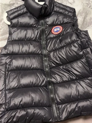 Svart dunväst från Canada Goose - Snygg svart dunväst från Canada Goose med klassisk logotyp på bröstet. Västen har hög krage, dragkedja framtill och quiltad design. Perfekt för lager-på-lager och håller dig varm med sin fluffiga dunfyllning. Passar till många olika stilar.