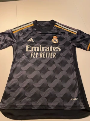Real Madrid bortatröja Adidas svart - Snygg svart bortatröja från Real Madrid med vågmönster, Adidas-logga och klubbmärke på bröstet. Tröjan har gula och vita detaljer på axlarna samt 'Emirates Fly Better' tryckt på framsidan. Tillverkad i lätt och ventilerande material med Aeroready-teknologi.