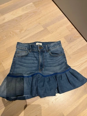 Egensydd Blå volangkjol  - Säljer en kort egensydd jeanskjol i blå denim med volangkant nedtill. Kjolen har klassiska fickor fram och bak, bälteshällor och knappstängning. Perfekt för dig som gillar en trendig och avslappnad stil. Frånbörjan var det ett par jeans från Lindex som jag sedan sydde om till kjol. 