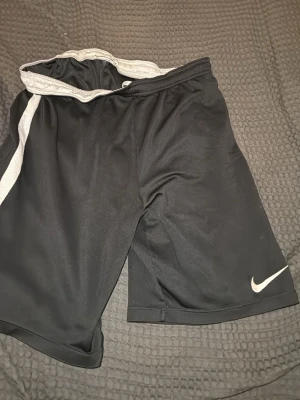 Svarta sportshorts från Nike - Säljer ett par svarta sportshorts från Nike med vit logga på vänster ben och grått innerfoder. Shortsen har elastisk midja och är tillverkade i ett lätt, snabbtorkande material som passar perfekt till träning eller chill.