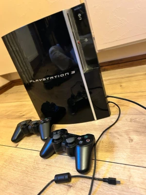 PlayStation 3 med två kontroller och gitarr - Säljer en svart PlayStation 3 från Sony med två trådlösa DualShock 3-kontroller och en svart Guitar Hero-gitarr. Konsolen har en snygg, blank finish och klassisk design. Perfekt för gamingkvällar och musikspel! Den har 19 spel inkluderande. 