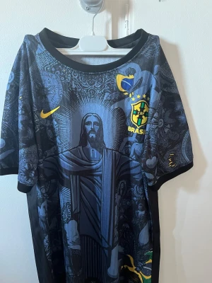 Nike Brasil svart fotbollströja - Snygg svart fotbollströja från Nike med Brasiliens landslagsmärke och gult Nike-logga. Tröjan har ett detaljerat grafiskt mönster med motiv av Kristusstatyn och andra symboler. Korta ärmar och rund hals. Perfekt för dig som älskar fotboll och streetwear. Inte äkta!