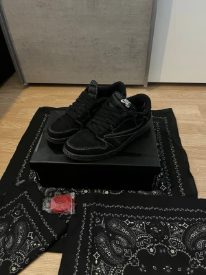 Nike SB Dunk Low x Travis Scott svart - Nike SB Dunk Low x Travis Scott i svart mocka med vita kontrastsömmar och svart sula. Klassisk Nike Swoosh på sidan och Travis Scott-detaljer. Skorna har svarta skosnören och extra röda snören medföljer. Perfekta för dig som gillar streetwear och unika samarbeten.