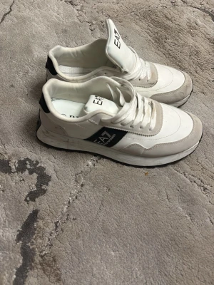 Vita EA7 Emporio Armani sneakers - Snygga vita sneakers från EA7 Emporio Armani med svarta detaljer och logga på sidan. Skorna har en klassisk låg siluett, vita snören och en kombination av läder och textilmaterial. Perfekta för en trendig och sportig look.