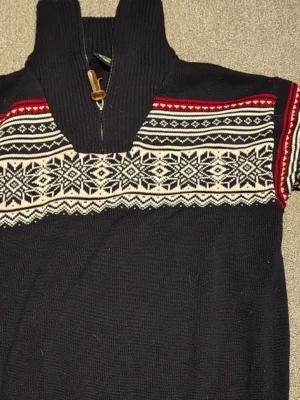 Mörkblå stickad tröja med mönster - Snygg mörkblå stickad tröja från Dale of Norway med klassiskt nordiskt mönster i vitt och rött över bröstet. Tröjan har hög krage med dragkedja och trädetalj. Perfekt för kalla dagar och ger en riktigt mysig vinterlook. Jätte skönt men har blivit för liten för mig 