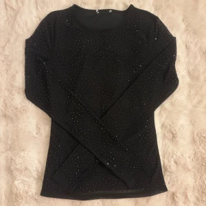 Svart topp med glittriga detaljer - Snygg svart långärmad topp med glittriga paljettdetaljer över hela tyget. Toppen har en rund halsringning och är figurnära i modellen. Perfekt för fest och utekvällar!