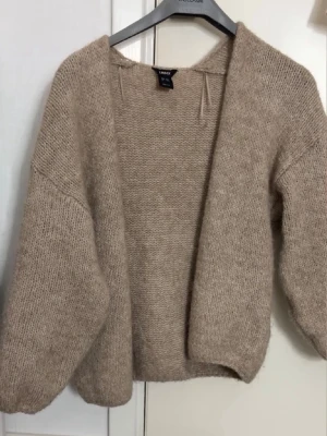 Beige stickad kofta från Lindex - Mysig beige stickad kofta från Lindex med öppen front och avslappnad passform. Koftan har långa ärmar och är tillverkad i ett mjukt, fluffigt material som känns skönt mot huden. Perfekt för lager på lager och enkel att matcha med olika outfits.