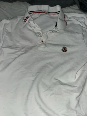 Vit Moncler pikétröja med logga - Klassisk vit pikétröja från Moncler med broderad logga på bröstet och snygga ränder i rött och blått på kragen. Tröjan har knappar framtill och är tillverkad i bomull för en skön och avslappnad stil. Pris kan diskuteras 
